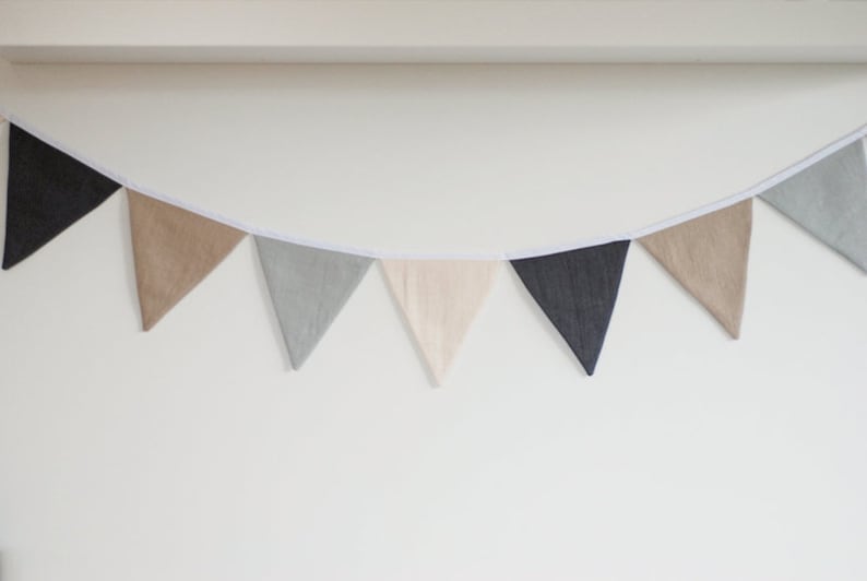 Puede incluir: Una guirnalda de banderines con siete banderas triangulares de tela en tonos alternos de gris carb&oacute;n, beige, gris claro y crema. Las banderas est&aacute;n unidas a una cinta de tela blanca continua, creando un adorno colgante decorativo.