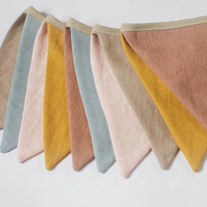 Può includere: Un banner di bandierine con otto bandierine triangolari in diverse tonalità di marrone, giallo, rosa e blu. Le bandierine sono realizzate in tessuto di lino e hanno un bordo color crema. Il banner è etichettato "JUMATA MADE".