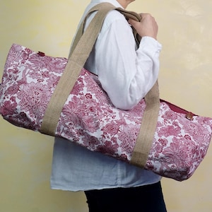 May include: A floral patterned tote bag with a white background and burgundy flowers. The bag has a rectangular shape with a zipper closure and a burlap shoulder strap. The bag is being carried over the shoulder.