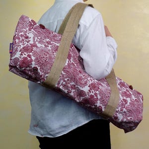 May include: A yoga mat bag with a floral pattern in shades of burgundy and white. The bag has a sturdy, natural-colored jute shoulder strap and trim. The bag is designed to carry a standard-sized yoga mat.