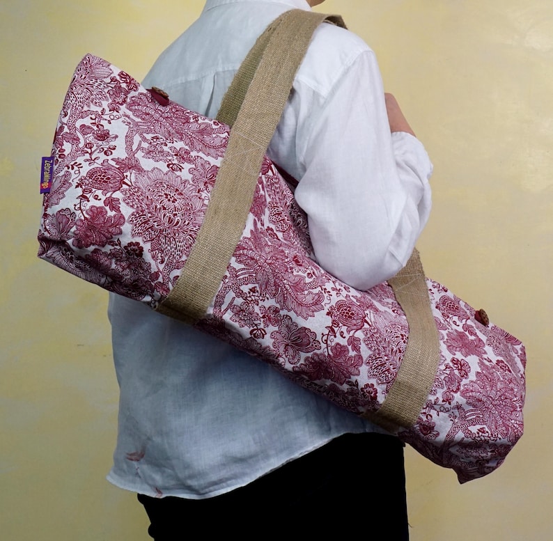 May include: A yoga mat bag with a floral pattern in shades of burgundy and white. The bag has a sturdy, natural-colored jute shoulder strap and trim. The bag is designed to carry a standard-sized yoga mat.