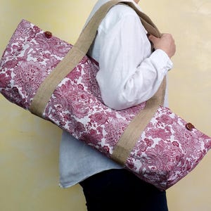 May include: A burgundy and white floral patterned yoga mat bag with a tan jute strap and trim. The bag has a button closure and is designed to carry a yoga mat. The bag is made of fabric.