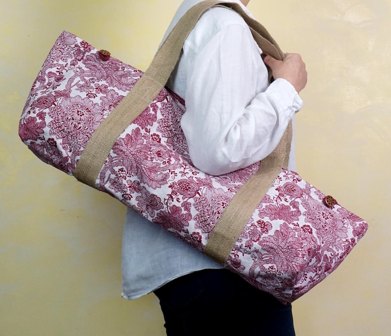 May include: A burgundy and white floral patterned yoga mat bag with a tan jute strap and trim. The bag has a button closure and is designed to carry a yoga mat. The bag is made of fabric.