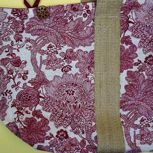 May include: A tote bag with a floral pattern in burgundy and white. The bag features a natural-colored jute strap and a wooden button closure. The floral design is detailed and covers the majority of the bag's surface.