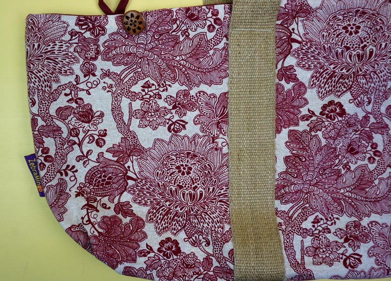 May include: A tote bag with a floral pattern in burgundy and white. The bag features a natural-colored jute strap and a wooden button closure. The floral design is detailed and covers the majority of the bag's surface.