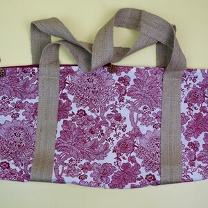May include: A tote bag with a floral pattern in shades of white and magenta. The bag has two burlap handles and is secured with wooden buttons and burgundy loops. The bag is designed for carrying items.