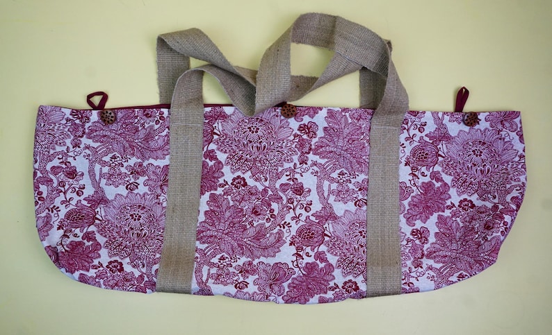 May include: A tote bag with a floral pattern in shades of white and magenta. The bag has two burlap handles and is secured with wooden buttons and burgundy loops. The bag is designed for carrying items.