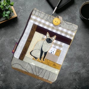 May include: A patchwork tablet case featuring a Siamese cat applique. The case is made of various fabrics in brown, beige, and gray tones, with a button closure. The brand name "LEORAMINGO" is visible on a small tag.