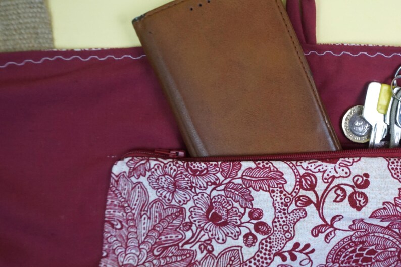 May include: A burgundy tote bag with a brown leather wallet partially visible. A patterned zippered pouch with a floral design in white and burgundy is in the front pocket. Keys and a coin are also visible.