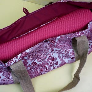 May include: A floral patterned yoga bag in shades of purple and white, with a rolled-up pink yoga mat inside. The bag has a burgundy interior and burlap handles. The bag is open, showing the contents.