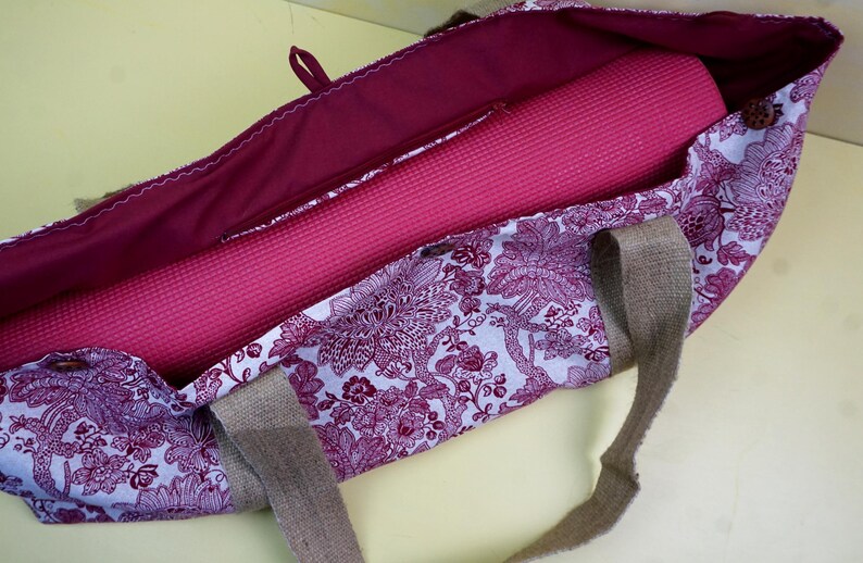 May include: A floral patterned yoga bag in shades of purple and white, with a rolled-up pink yoga mat inside. The bag has a burgundy interior and burlap handles. The bag is open, showing the contents.