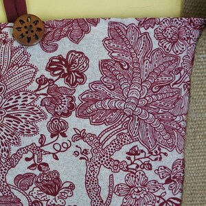 May include: Close-up of a fabric with a floral pattern in burgundy and white. A wooden button is attached to the top. The fabric is part of a larger item, possibly a tote bag or similar accessory.