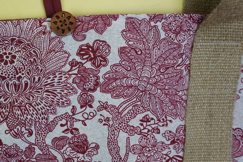 May include: Close-up of a fabric with a floral pattern in burgundy and white. A wooden button is attached to the top. The fabric is part of a larger item, possibly a tote bag or similar accessory.