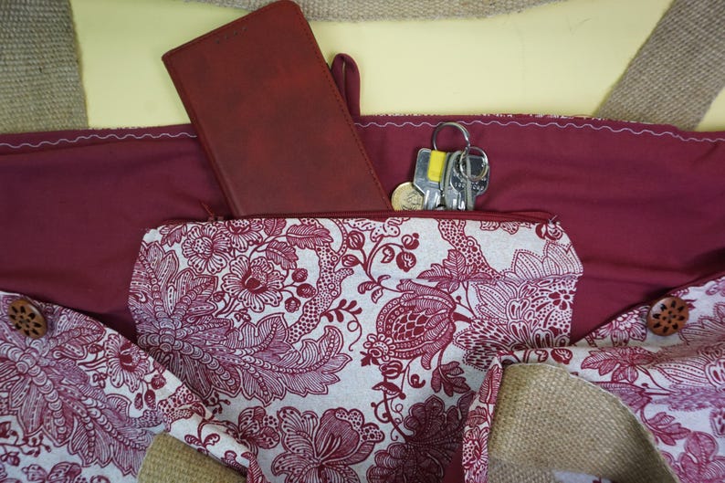 May include: Burgundy tote bag with a floral patterned pocket. A matching burgundy wallet and a set of keys are visible inside the bag. The bag has wooden button closures and a neutral-colored burlap trim.