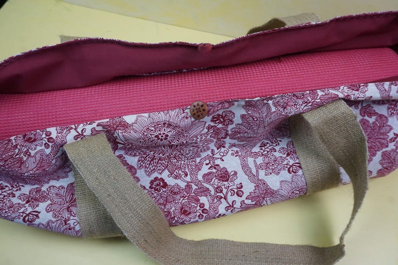 May include: A floral print yoga mat bag with a matching yoga mat inside. The bag features a white background with a burgundy floral pattern, burgundy lining, and tan handles. A wooden button secures the bag.
