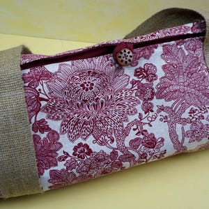 May include: A cylindrical bag with a floral pattern in burgundy and white. The bag has sturdy, tan-colored burlap handles and a wooden button closure. The floral design is a detailed, stylized pattern.