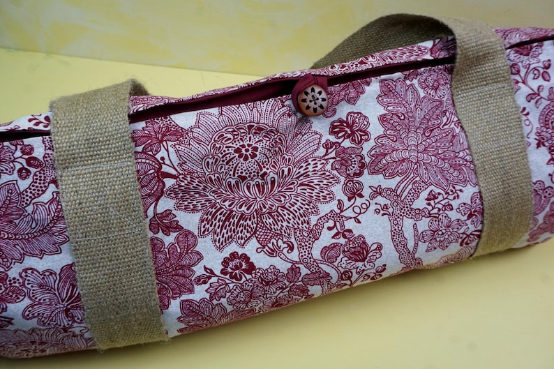 May include: A cylindrical bag with a floral pattern in burgundy and white. The bag has sturdy, tan-colored burlap handles and a wooden button closure. The floral design is a detailed, stylized pattern.