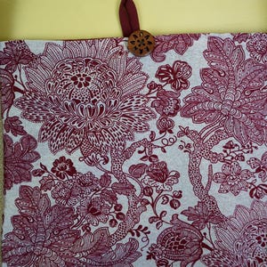 May include: Tote bag with a floral pattern in burgundy and white. The bag has a burlap-style strap and a wooden button closure. The floral design features large, detailed blooms and foliage.