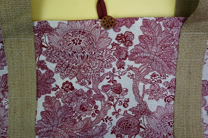 May include: Tote bag with a floral pattern in burgundy and white. The bag has a burlap-style strap and a wooden button closure. The floral design features large, detailed blooms and foliage.