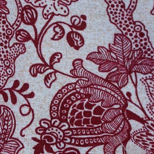 May include: Close-up of a fabric with a cream-colored background and a repeating pattern of dark red floral and botanical elements. The design includes stylized flowers, leaves, and branches, creating a textured, decorative surface.