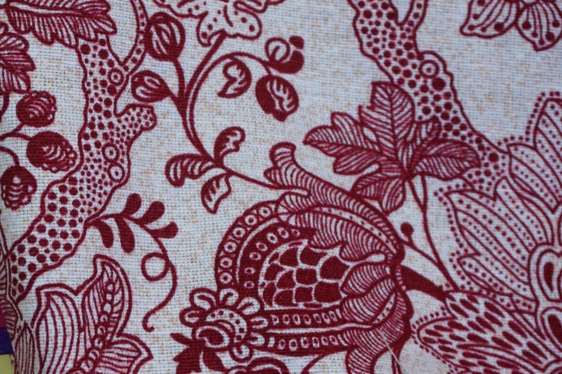 May include: Close-up of a fabric with a cream-colored background and a repeating pattern of dark red floral and botanical elements. The design includes stylized flowers, leaves, and branches, creating a textured, decorative surface.