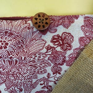 May include: Close-up of a fabric with a floral pattern in burgundy and white. A wooden button with a floral design is attached to the fabric. A section of tan burlap fabric is visible.