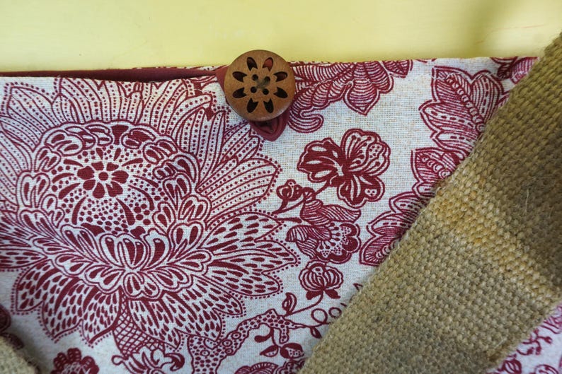 May include: Close-up of a fabric with a floral pattern in burgundy and white. A wooden button with a floral design is attached to the fabric. A section of tan burlap fabric is visible.