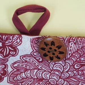 May include: Close-up of a fabric item with a floral pattern in white and burgundy. A burgundy loop handle is attached to the top. A wooden button with a floral design is sewn onto the fabric.