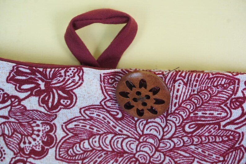 May include: Close-up of a fabric item with a floral pattern in white and burgundy. A burgundy loop handle is attached to the top. A wooden button with a floral design is sewn onto the fabric.