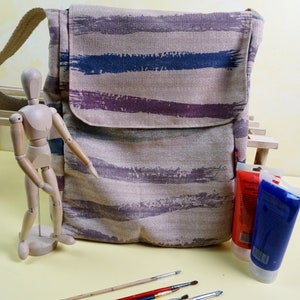 May include: A beige canvas messenger bag with horizontal brushstroke stripes in shades of purple and blue. The bag has a flap closure and a long strap. Paint tubes, paintbrushes, and a wooden artist's mannequin are also visible.