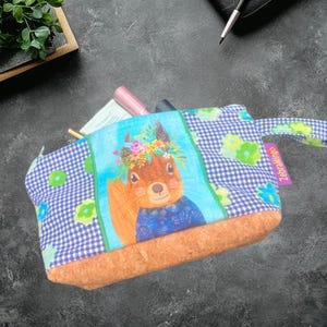 May include: A small, rectangular cosmetic bag with a blue and white checkered pattern and a cork base. The bag features a cartoon squirrel wearing a flower crown. The brand name "ZebraMingo" is printed on the side.