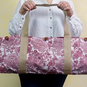May include: A large tote bag with a floral pattern in shades of red and white. The bag has sturdy burlap handles and is secured with wooden buttons. The bag is designed for carrying items.