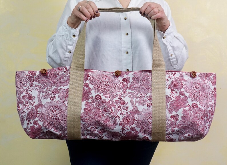 May include: A large tote bag with a floral pattern in shades of red and white. The bag has sturdy burlap handles and is secured with wooden buttons. The bag is designed for carrying items.