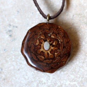 May include: A brown wood pendant with a white opal stone in the center. The pendant is surrounded by a detailed wood design and is attached to a brown leather cord.