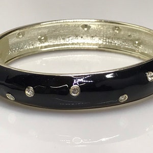 Puede incluir: Un brazalete de esmalte negro con detalles plateados y brillantes transparentes.
