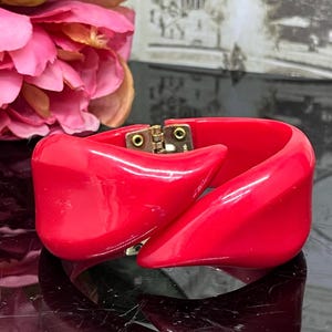 Puede incluir: Una pulsera roja vibrante y escultural con un cierre dorado. La pulsera tiene un diseño curvo y geométrico. El cierre es rectangular con dos tornillos pequeños. Una pieza de declaración audaz.