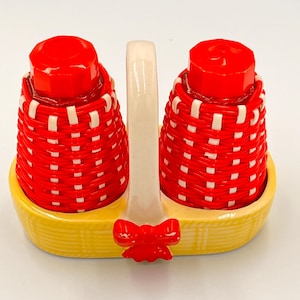 Può includere: Un set vintage di saliera e pepiera. I shaker rossi hanno un design a cestino intrecciato e coperchi rossi. Sono in un supporto giallo a forma di cestino con un manico bianco e un fiocco rosso.