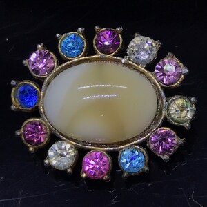 May include: A white oval cabochon surrounded by a ring of colorful rhinestones. The rhinestones are in shades of pink, blue, and clear.