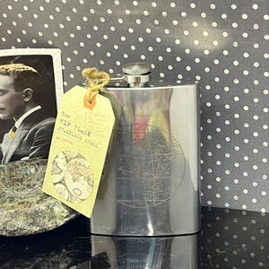 Könnte beinhalten: Eine Edelstahl-Hüftflasche mit Kartenmuster, Fassungsvermögen ca. 237 ml. Ein Anhänger mit der Aufschrift "HIP FLASK STAINLESS STEEL" ist angebracht. Die Flasche hat einen silbernen Deckel und wird vor einem gepunkteten Hintergrund präsentiert.