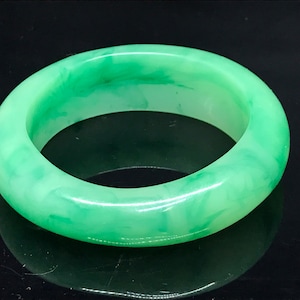 Könnte beinhalten: Ein Jade-Armreif. Das Armband hat eine hellgrüne Farbe mit marmoriertem Aussehen. Die glatte, polierte Oberfläche reflektiert das Licht und hebt die natürlichen Variationen des Steins hervor. Dieses Schmuckstück ist ein klassisches Accessoire.