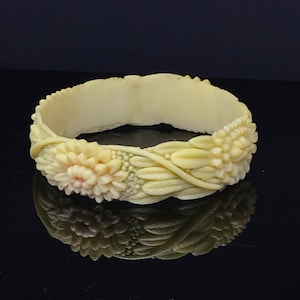 Puede incluir: Brazalete rígido de color marfil con diseño floral tallado. El brazalete presenta tallados detallados de flores y hojas, con una superficie ligeramente texturizada. Las flores tienen un toque de rosa en el centro, añadiendo un contraste sutil.