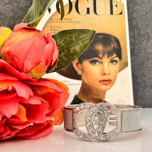 Peut inclure: Un bracelet argenté avec un motif double-C incrusté de cristaux transparents. Le bracelet a des sections rouges et argentées scintillantes. Une couverture vintage du magazine Vogue et une fleur rouge en arrière-plan.