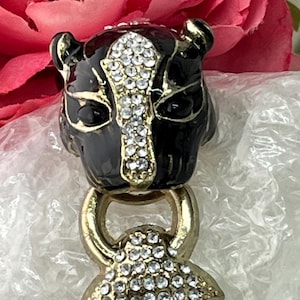 Peut inclure: Un bouchon de bouteille noir et doré avec un motif de tête de panthère. La tête de la panthère est ornée de strass étincelants, et le bouchon a un anneau et une base dorés. Des fleurs roses sont en arrière-plan.