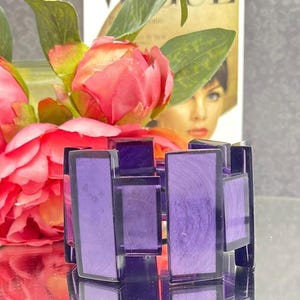 Puede incluir: Un brazalete geométrico púrpura con formas rectangulares y cuadradas. El brazalete tiene un marco púrpura oscuro y piezas centrales púrpuras más claras. El brazalete se exhibe frente a flores rosas y una revista.