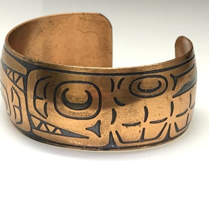 May include: A copper bracelet with a black etched design of stylised geometric shapes. The bracelet is open and has a rounded shape.