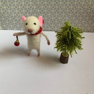 May include: A white felt mouse wearing a red scarf and holding a red ornament stands next to a small green felt Christmas tree.