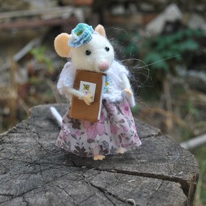 May include: A white felt mouse wearing a floral dress and a blue flower crown holds a small brown book. The mouse is standing on a tree stump.