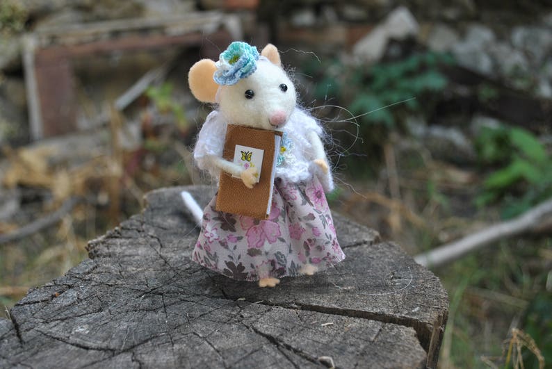 May include: A white felt mouse wearing a floral dress and a blue flower crown holds a small brown book. The mouse is standing on a tree stump.