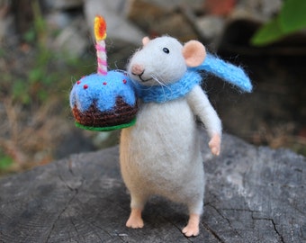 Può includere: Un topo bianco di feltro con una sciarpa blu tiene una torta di compleanno blu e marrone con una candela accesa.
