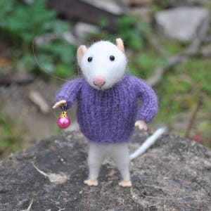 May include: A handcrafted felt mouse figurine wearing a purple sweater and holding a pink ornament. The mouse has a white body, gray legs, and small ears. The ornament is pink with gold accents. The figurine is standing on a tree stump.